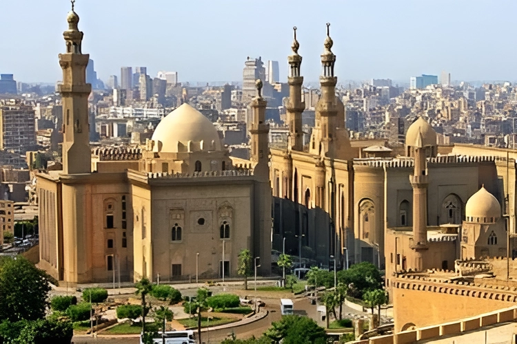 The image shows the historic Sultan Hassan Mosque and Madrasa in Cairo, with its large dome and minarets set against a backdrop of the city skyline.