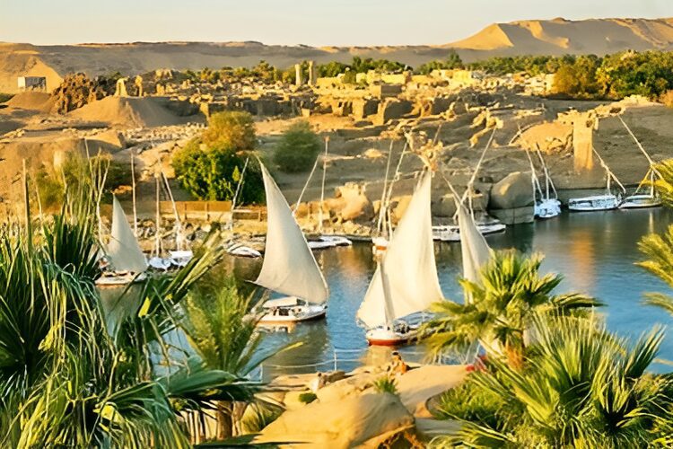 Sailboats with white sails glide on a river surrounded by lush greenery and ancient ruins under a clear sky.