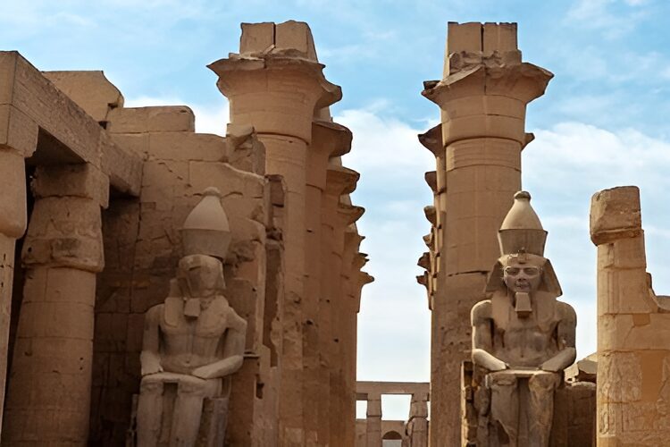 Two large ancient stone statues are seated amidst towering columns, set against a clear sky.