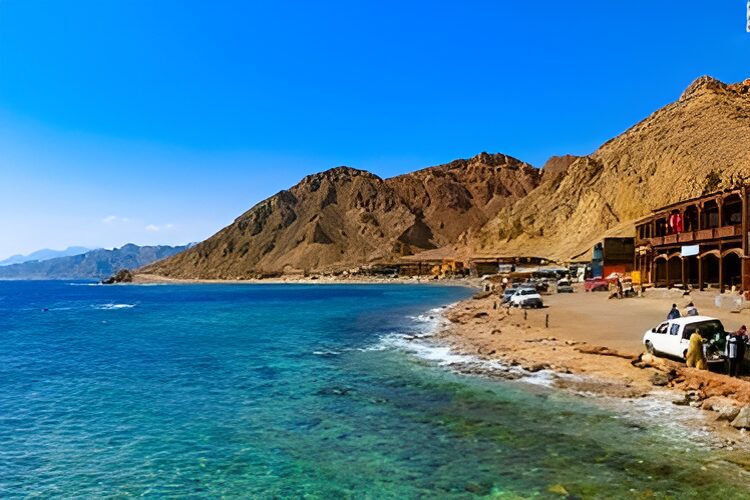 Coastal view of a rocky shoreline with clear blue water, cars parked along the edge, and brown mountains in the background under a clear blue sky.