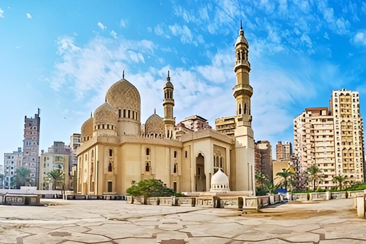 A large mosque with beige domes and minarets stands under a blue sky, surrounded by tall buildings.