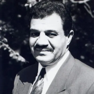 A man in a suit and tie stands outdoors, smiling slightly. The photo is black and white with trees in the background.