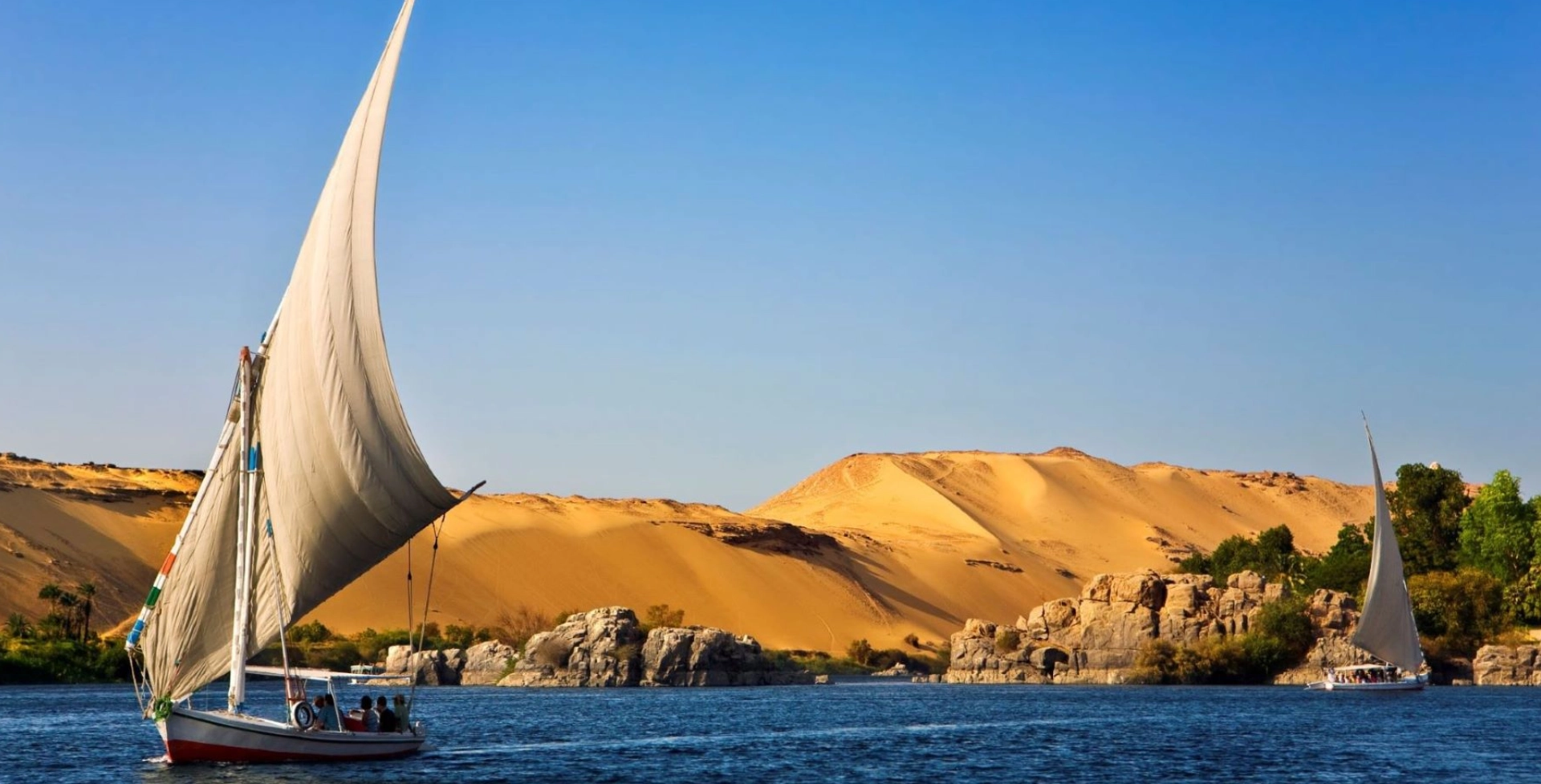 A sailboat glides across a river with a backdrop of sandy dunes and rocky terrain under a clear blue sky.