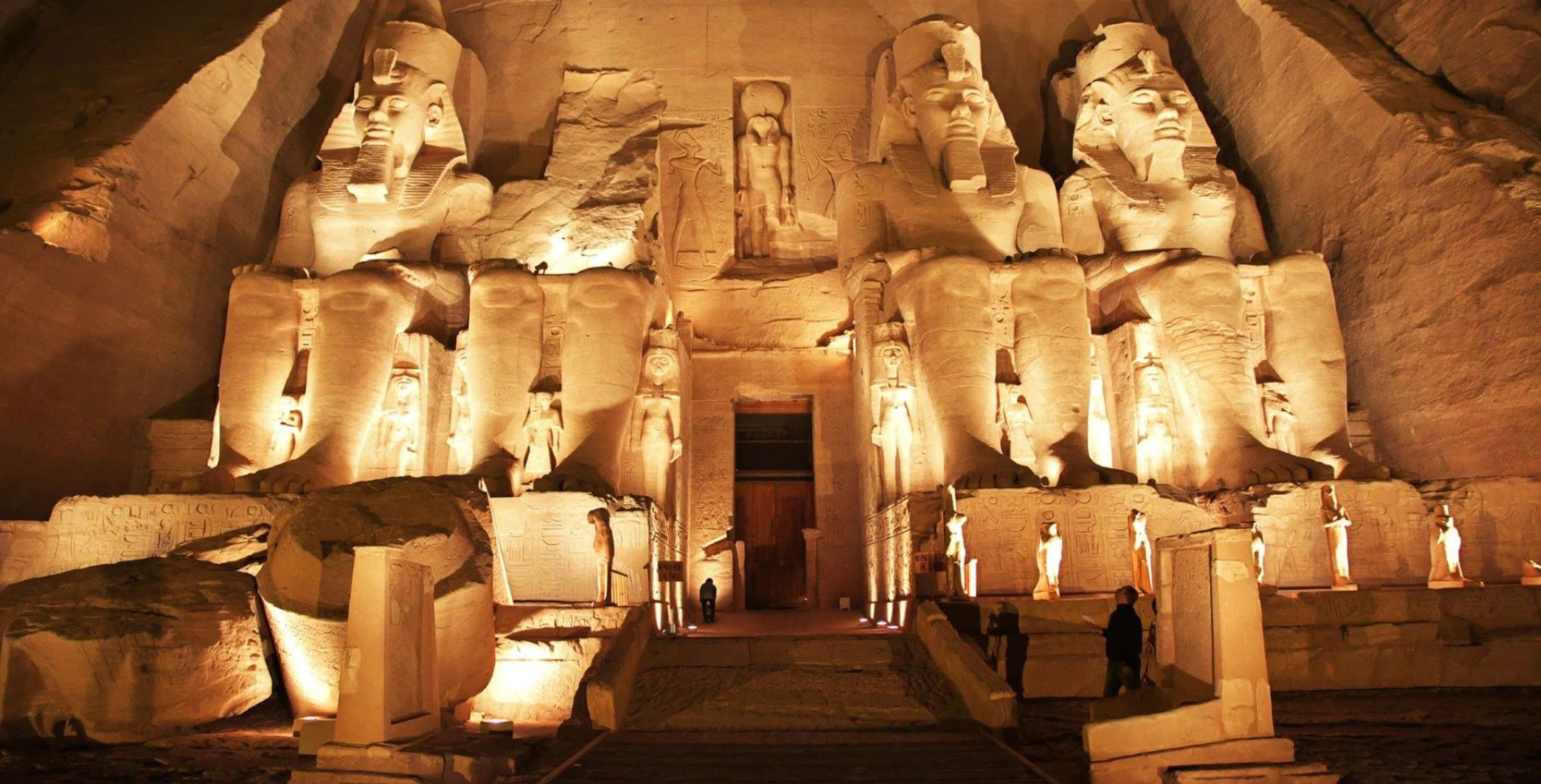 Illuminated ancient temple façade with four large seated statues and towering pillars at night.