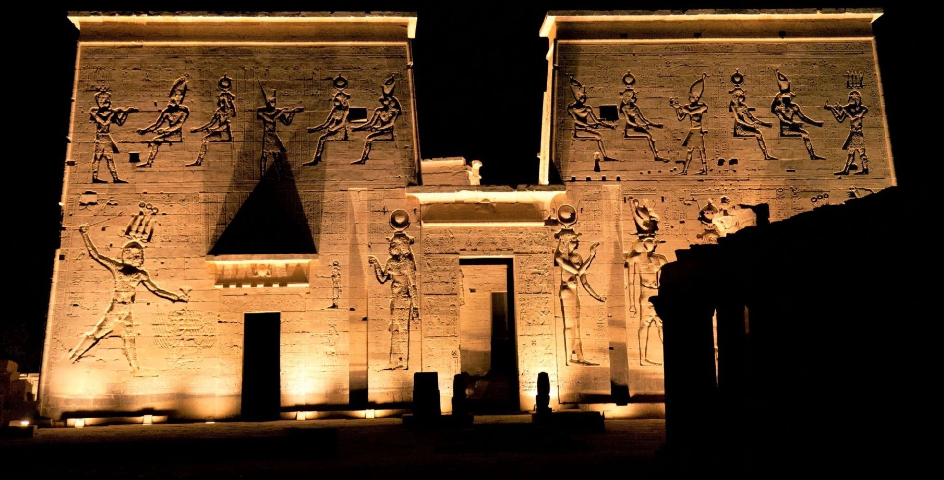 Night view of an illuminated ancient Egyptian temple with detailed carvings and hieroglyphics on the walls.