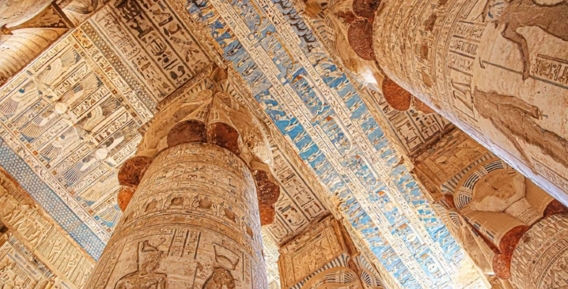 Ancient Egyptian temple ceiling with detailed carvings and paintings in blue and gold hues, supported by large decorated columns featuring hieroglyphics and figures.