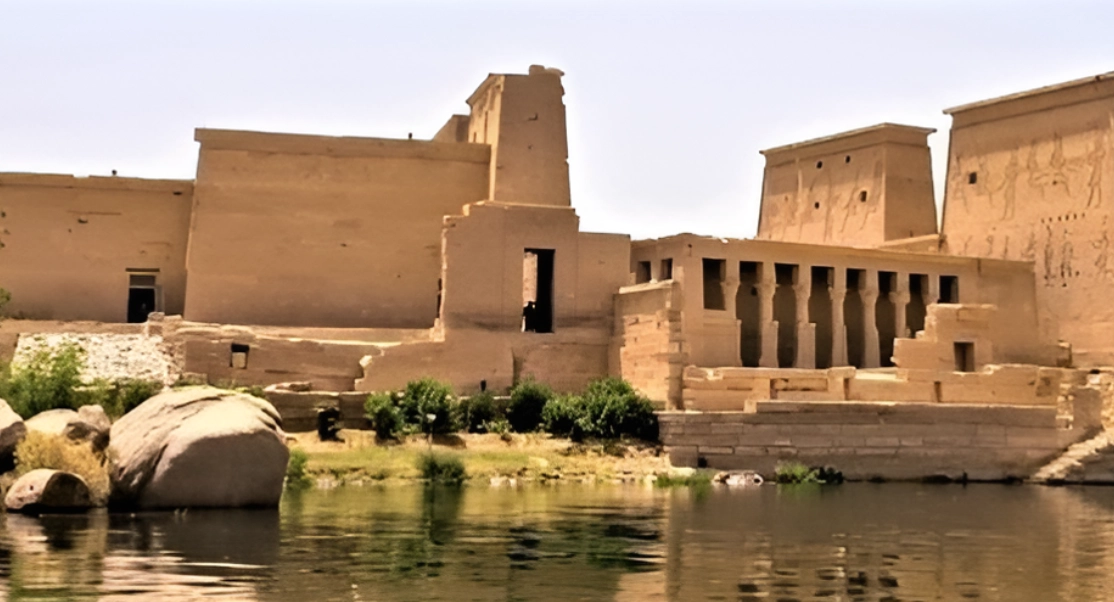 Ancient stone temple complex with large columns and a calm river in the foreground, under a clear sky.