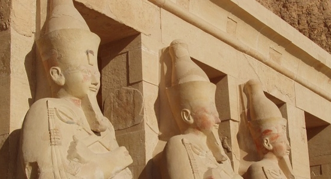 Ancient Egyptian statues of pharaohs with tall headdresses carved into a stone wall, partially lit by sunlight.