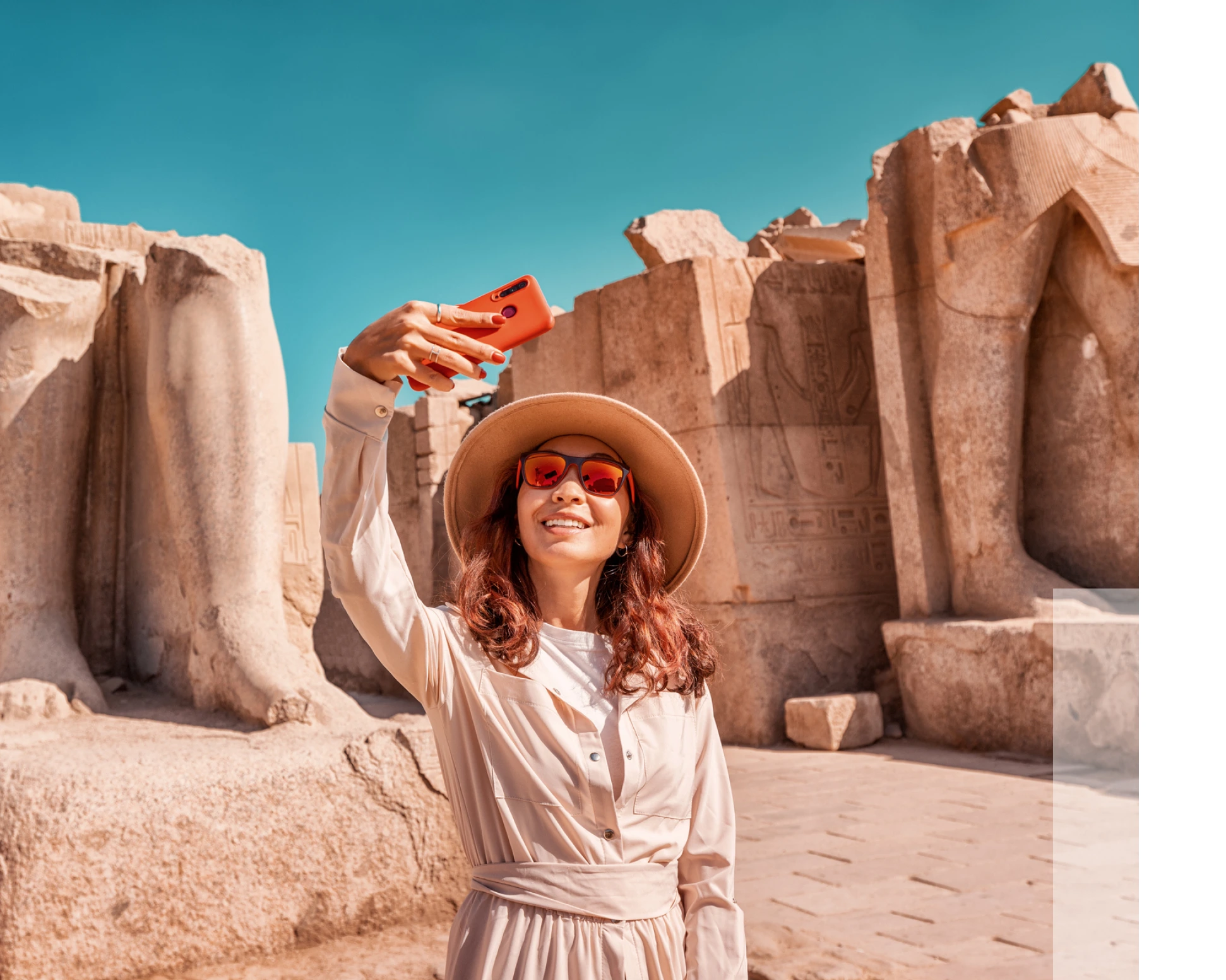 A woman in a hat and sunglasses takes a selfie with a smartphone in front of ancient stone ruins under a blue sky.