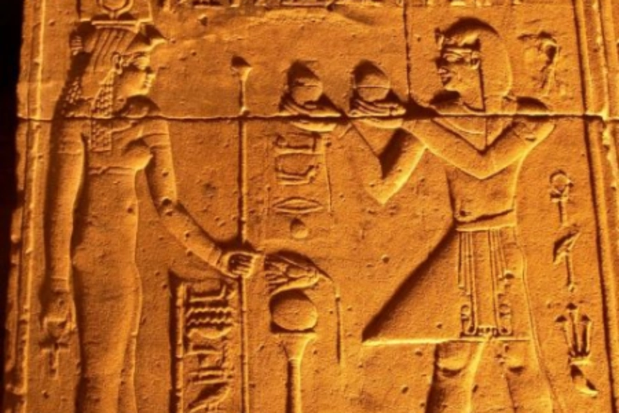 Ancient Egyptian wall carving depicting two figures with hieroglyphs, one holding an offering in raised hands.